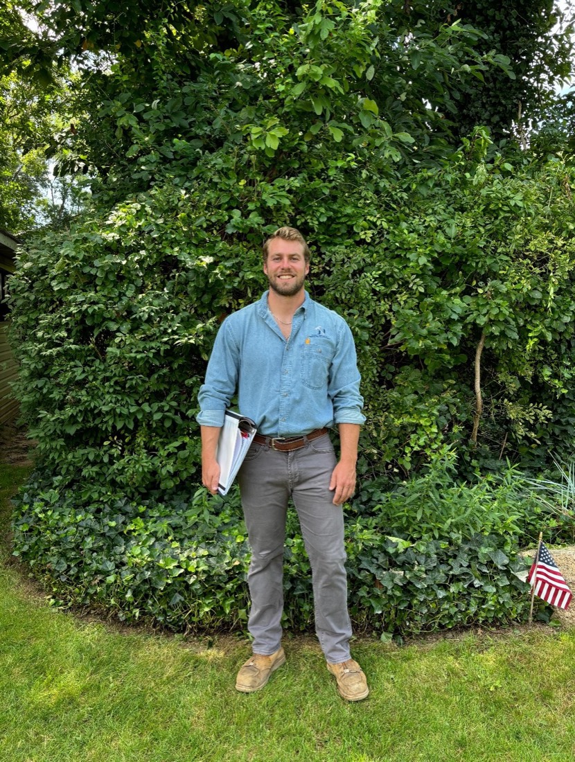 The owner of Nielsen Services & Property Management, in a work shirt with an inspection clipboard beside a client's home and an American flag.